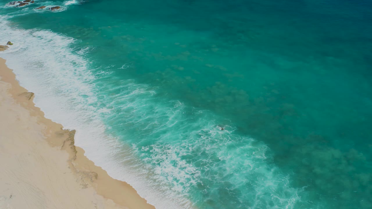 Aerial view of El Cardoncito shoreline with more beach visible at left, bright foam curling over turquoise shallows, and a small dark person or object near mid-frame water.