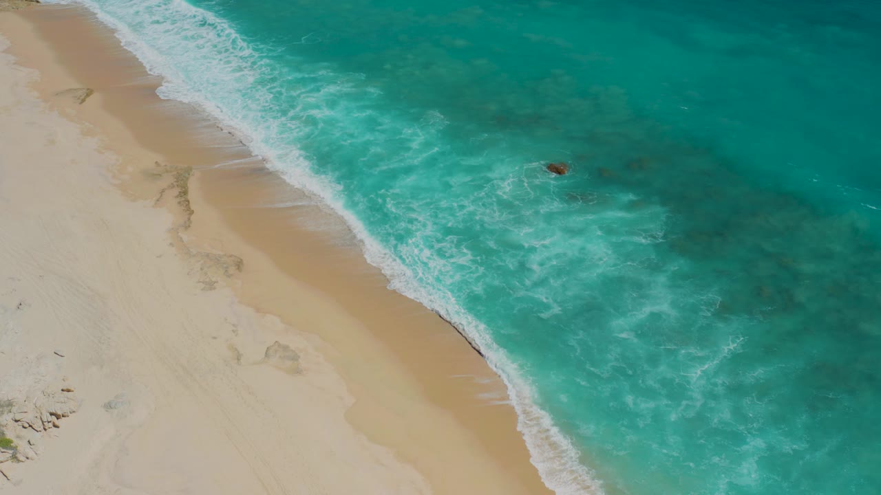 High aerial view in El Cardoncito, Baja California Sur, with pale sand on the left, turquoise water on the right, and a small dark object or distant person near the breaking surf.