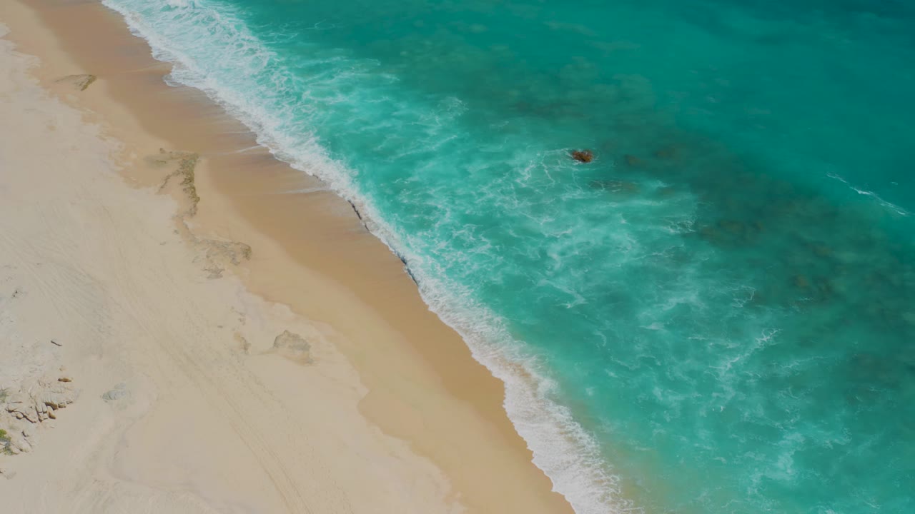 The El Cardoncito shoreline fills the frame diagonally, with white wash spreading across the sand and the small dark object or distant person still visible beyond the shore break.