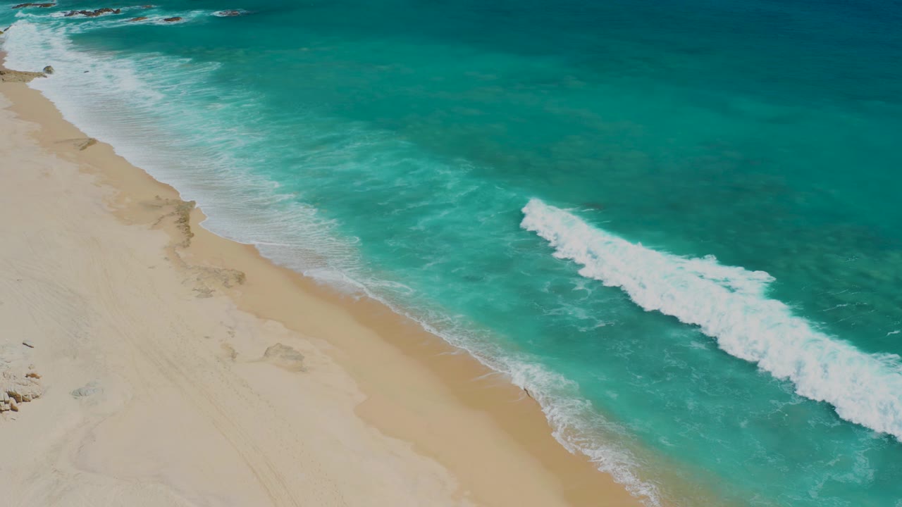 Aerial frame over El Cardoncito, Baja California Sur, showing pale sand at left, turquoise water at right, a white breaking wave offshore, and no visible people.