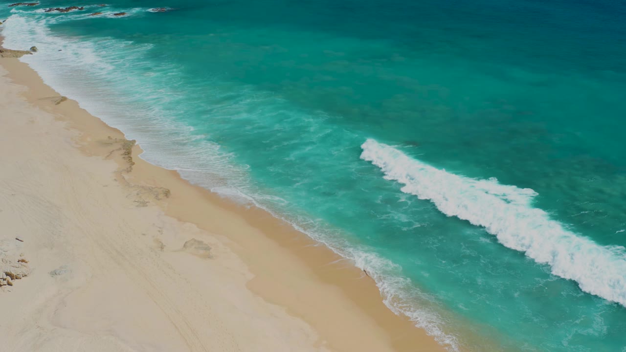 Aerial view at El Cardoncito with the beach still along the left edge, a foamy wave advancing through the center-right water, and no visible people on the sand.