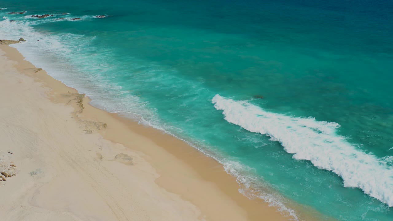 Aerial frame from El Cardoncito showing a larger white wave patch in the right half, pale sand on the left, darker offshore water above, and no visible people.