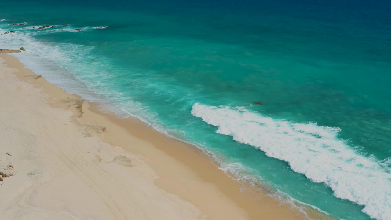 Aerial view of El Cardoncito with the surf line spread farther across the lower right, tan sand along the left side, and no visible people or built structures.