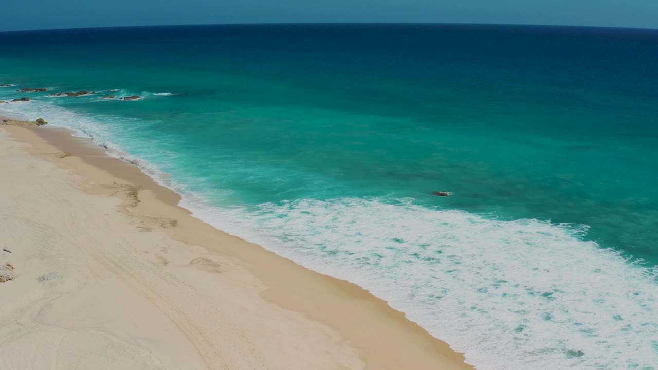 Aerial frame from El Cardoncito, Baja California Sur, Mexico, showing turquoise water rolling onto a pale sandy beach with darker ocean near the horizon and no visible people.