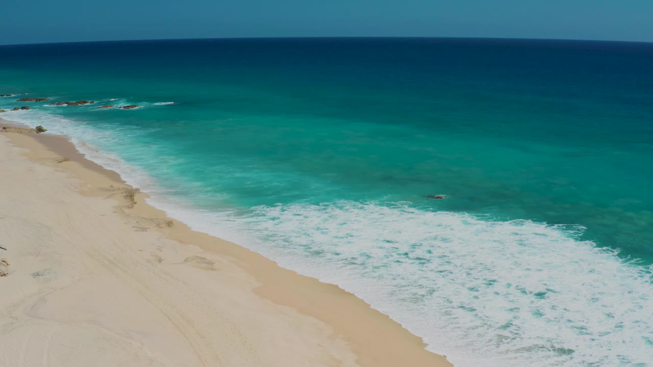 Aerial frame from El Cardoncito showing a diagonal sand edge, foamy surf across the center, clear turquoise water, deeper blue ocean at the top, and no visible people.