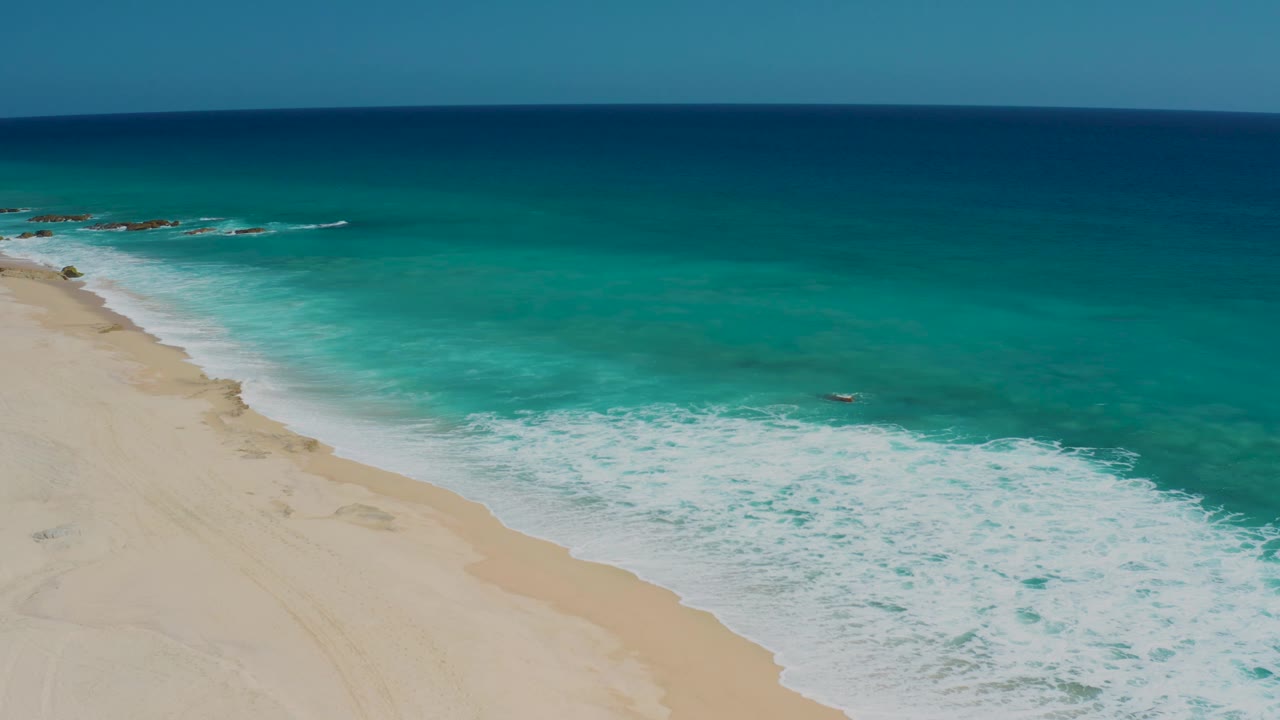 Elevated view over El Cardoncito with pale sand along the left, a broad band of breaking white surf, open turquoise water, and no visible people on the beach.