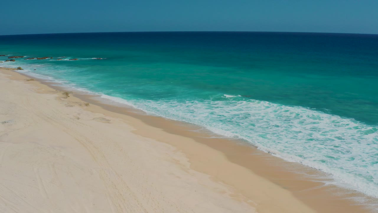Aerial view of El Cardoncito shoreline with pale sand filling the lower frame, turquoise surf breaking diagonally, and no clearly visible people.
