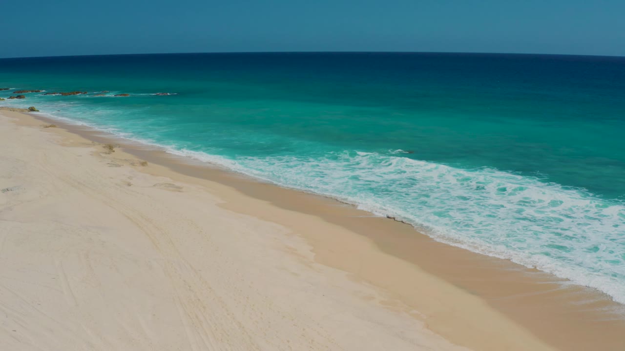 El Cardoncito shoreline seen from above, with a smooth beige beach, a thin wet-sand edge, turquoise water, and no clearly visible people.
