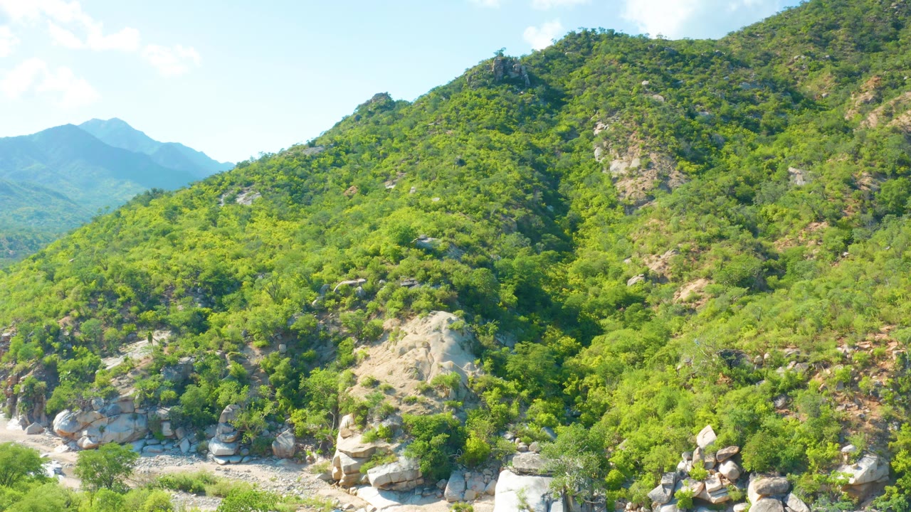 A vegetated mountain slope in La Palma, Baja California Sur, Mexico is framed with boulders near the base, hazy blue mountains behind, and no visible people.