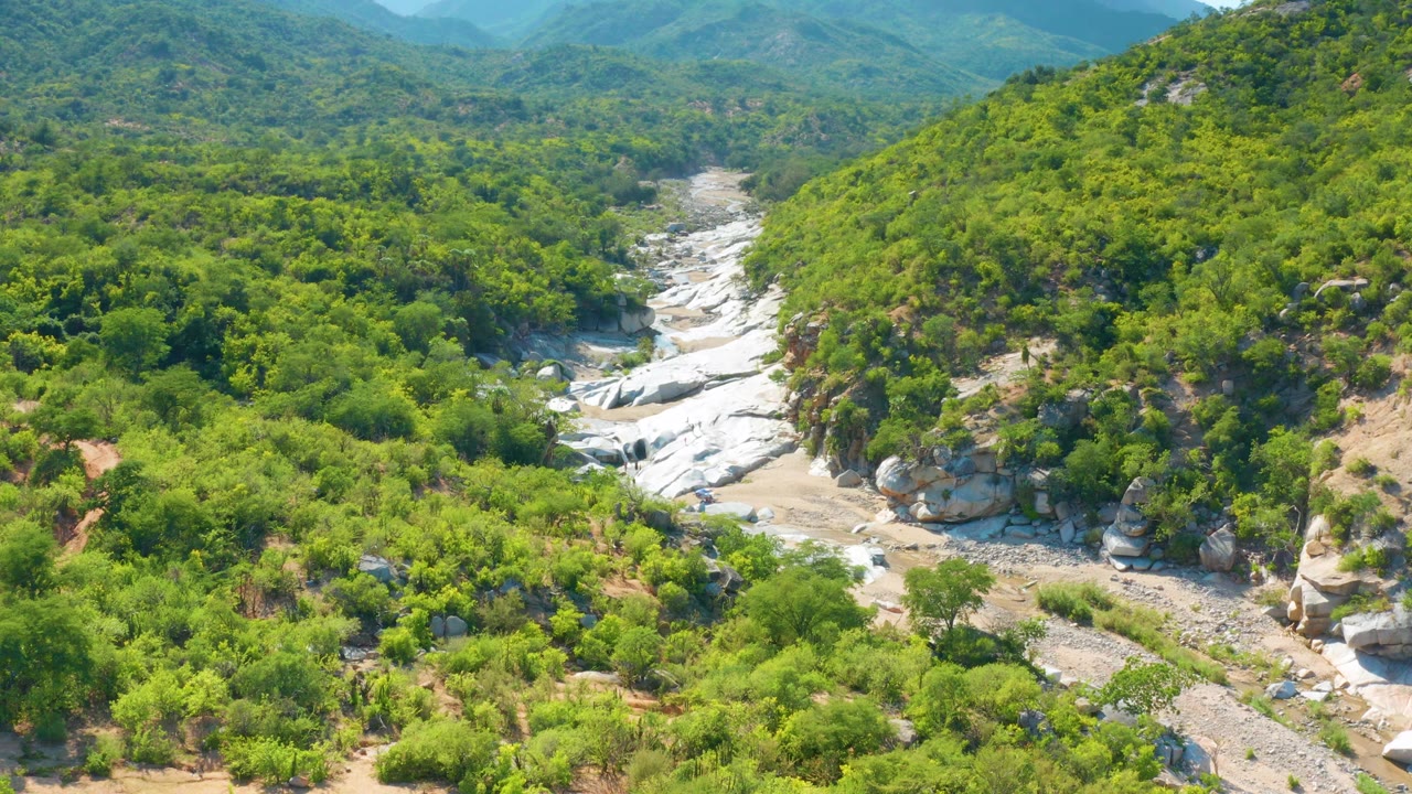 Aerial frame from La Palma, Baja California Sur with the rocky wash centered between brush-covered slopes and a hazy mountain background, with no visible people.