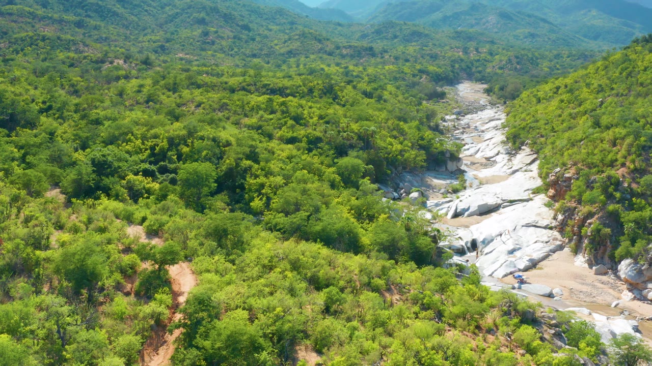 The La Palma aerial view shows a rocky valley floor cutting through dense brush and low trees, with layered green mountains fading into haze and no visible people.
