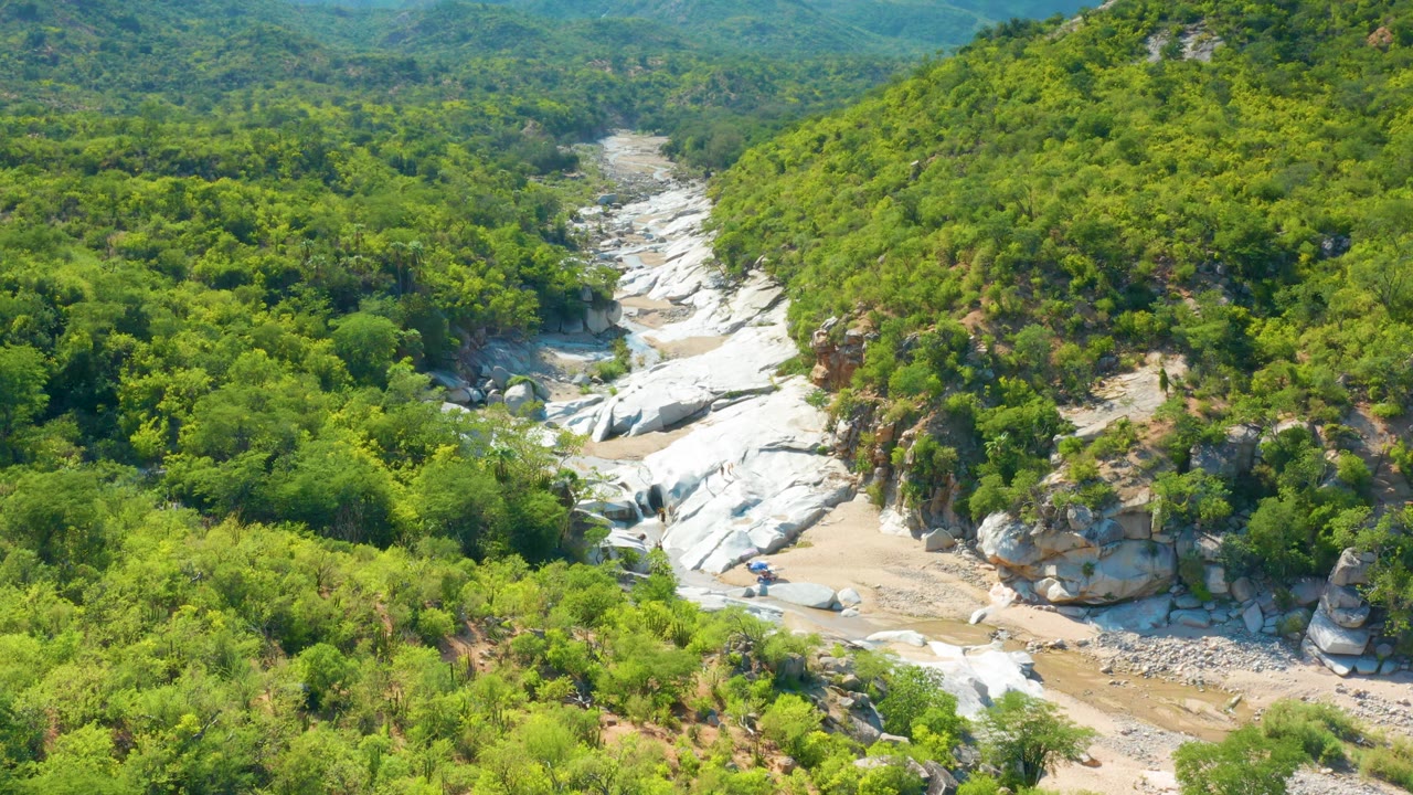 Aerial frame in La Palma, Baja California Sur, showing a winding dry wash bordered by white rock outcrops and thick green vegetation, with no visible people.