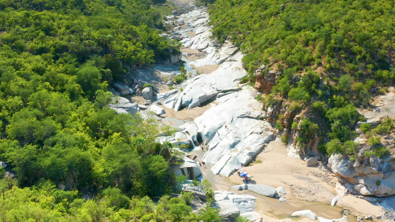 High aerial frame over La Palma, Baja California Sur, showing the rocky wash centered between dense vegetation and a reddish exposed slope, with no visible people.