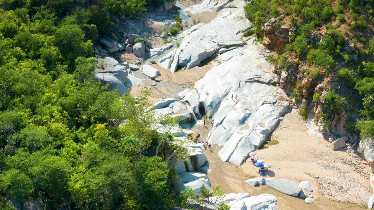 High aerial frame in La Palma, Baja California Sur, with large pale rock surfaces cutting through green vegetation and tan sand, with no visible people.