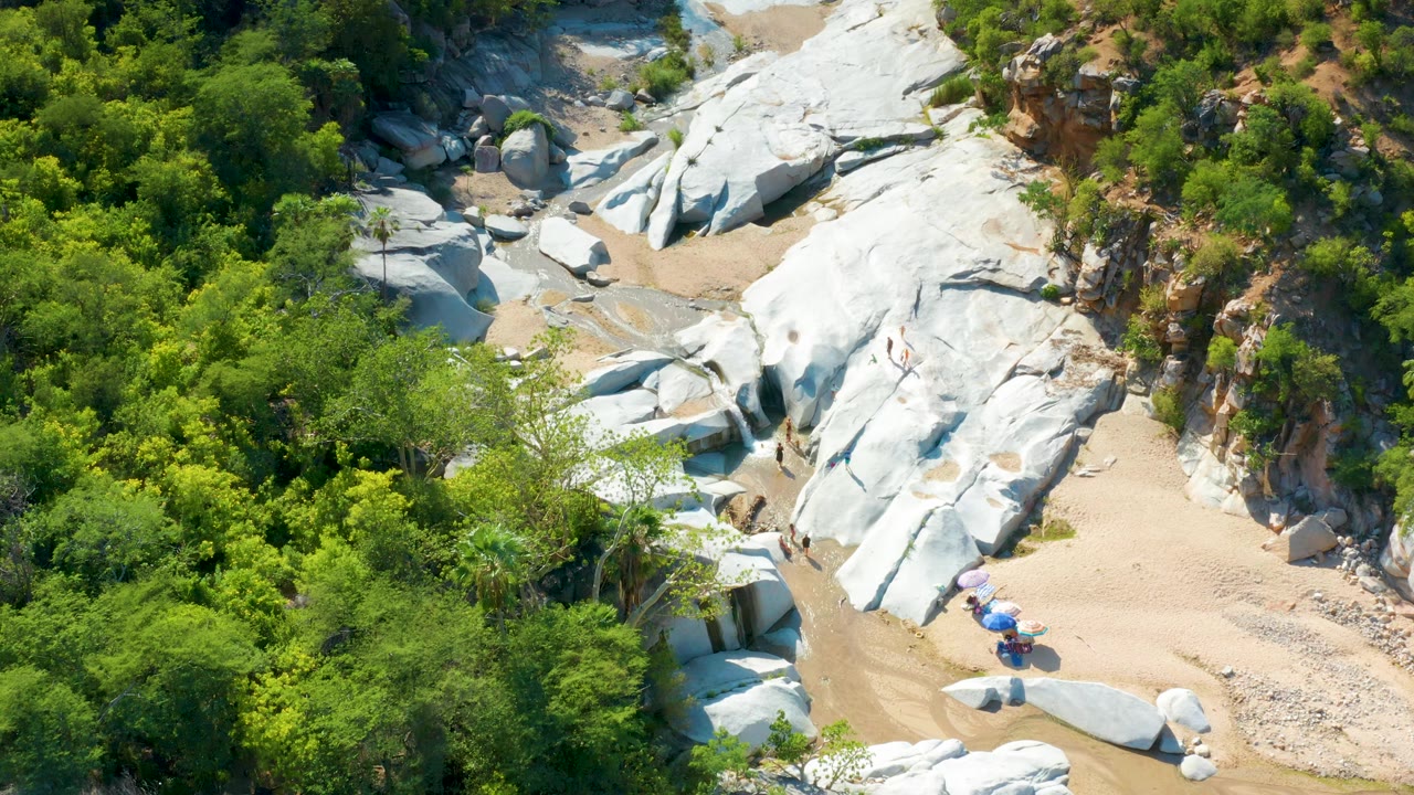 Top-down aerial view in La Palma, Baja California Sur, showing smooth white boulders, narrow sandy channels, and thick green foliage, with no visible people.