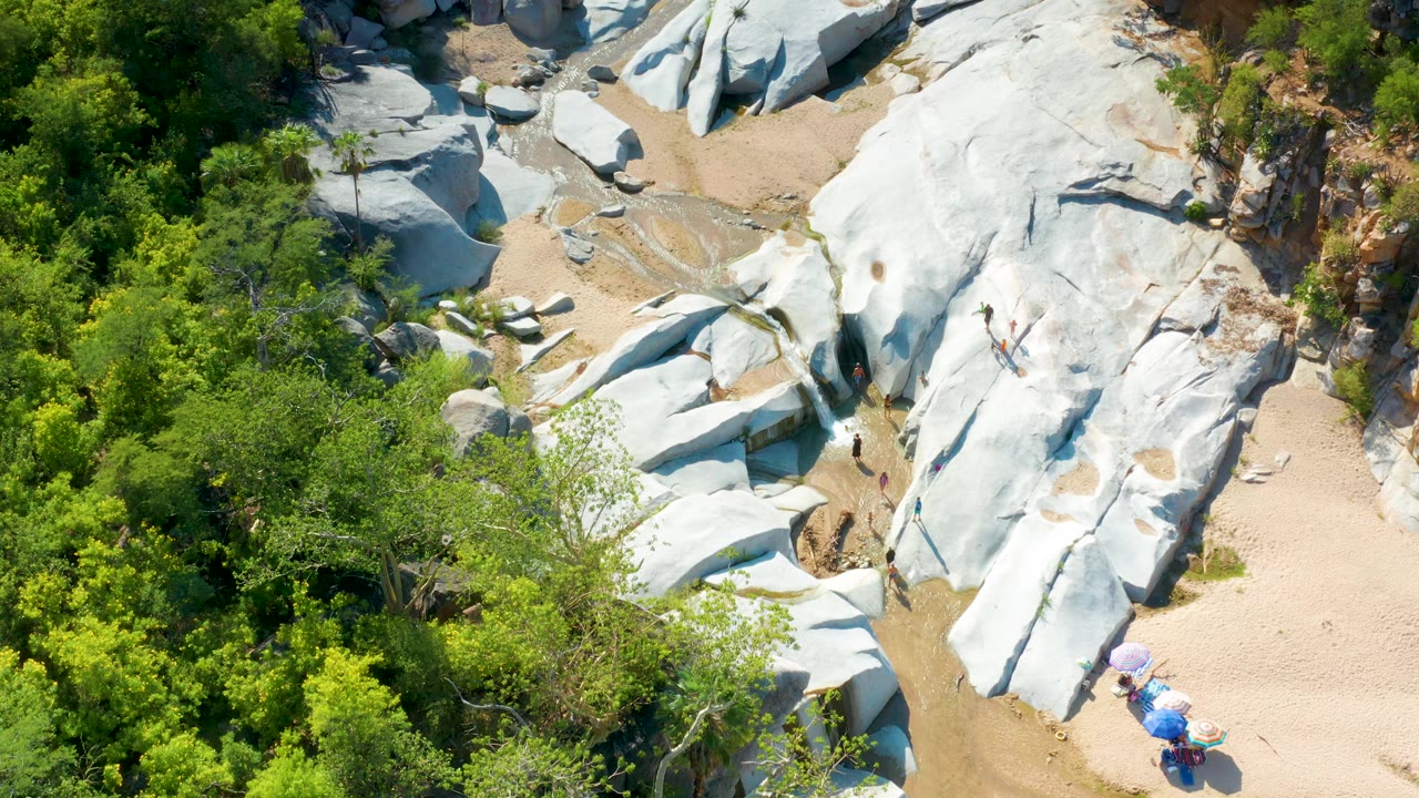 Aerial frame over La Palma, Baja California Sur with the creek visible between pale rock slabs, green canopy on the left, sandy wash areas, and no visible people.