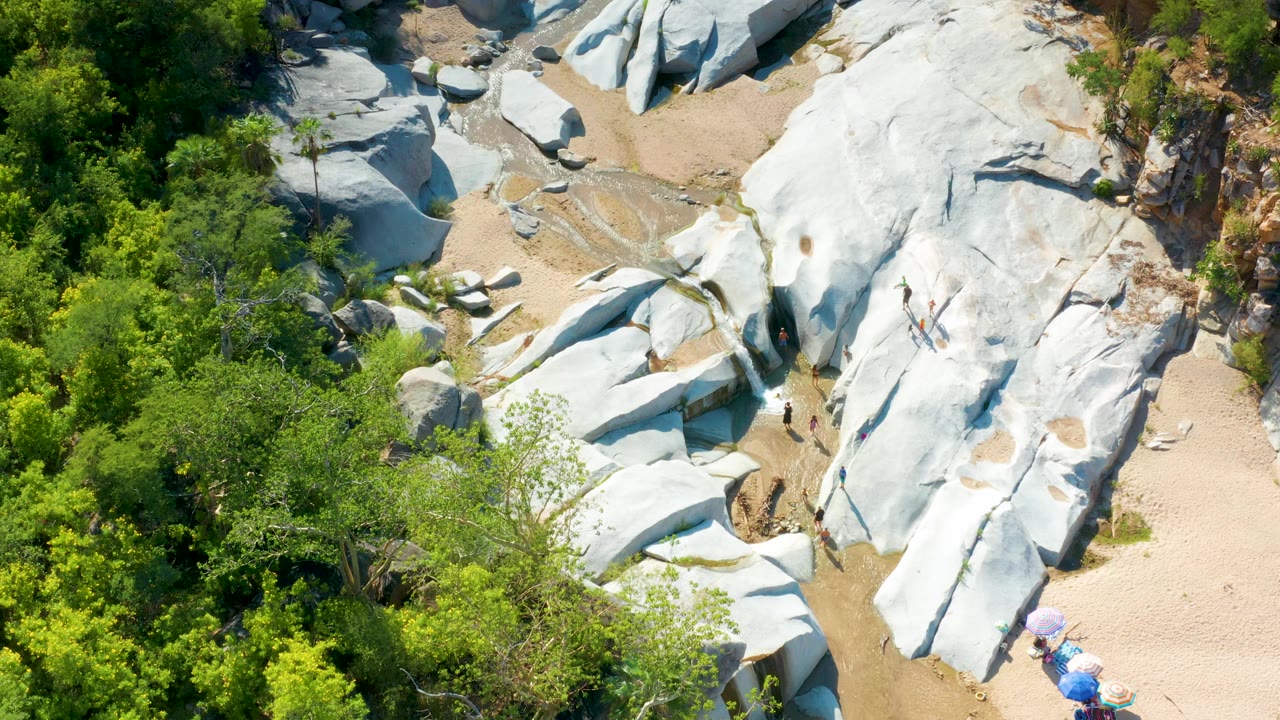 Top-down La Palma, Baja California Sur frame with dense trees, white boulders, a shallow creek bed, sandy ground, small blue and purple umbrellas near the bottom edge, and no visible people.