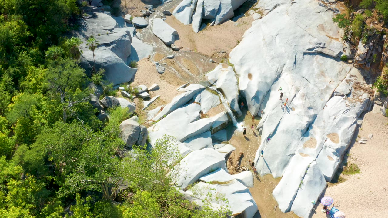 Aerial view in La Palma, Baja California Sur showing a creek bend through sand and pale boulders, shaded green vegetation on the left, bright rock surfaces, and no visible people.