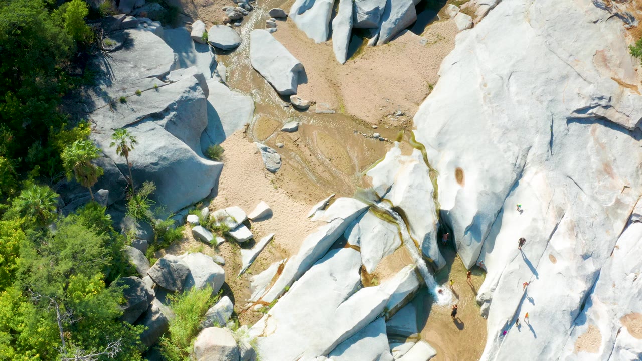 Top-down La Palma frame showing pale stone shapes crossing the right side, tan sand in the middle, a narrow wet channel, tree canopy at left, and no visible people.