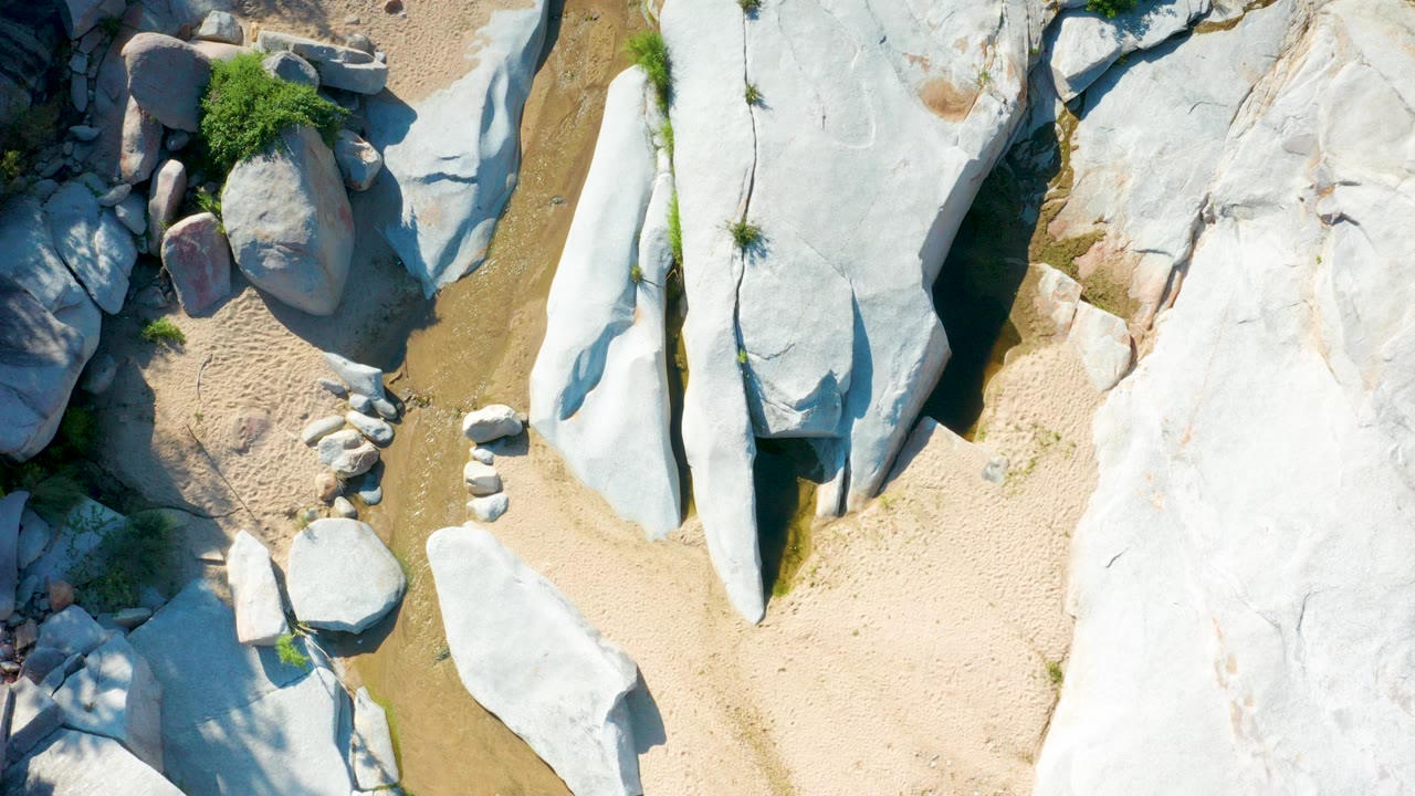 Overhead frame from La Palma, Baja California Sur showing pale rock slabs on the right, tan sand below, shallow brown-green water at left, scattered shrubs, and no visible people.