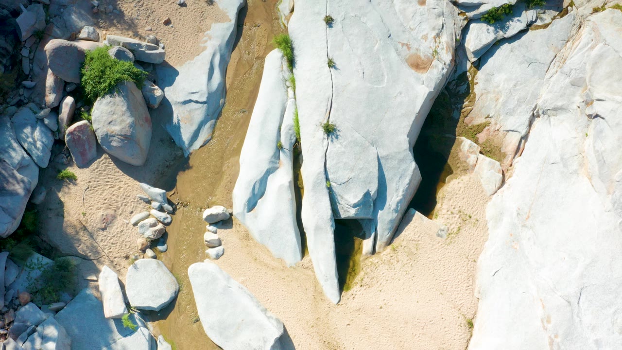 Overhead La Palma, Baja California Sur frame showing pale rock filling the right side, a sandy wash through the center, shallow water along the rocks, shrubs, and no visible people.