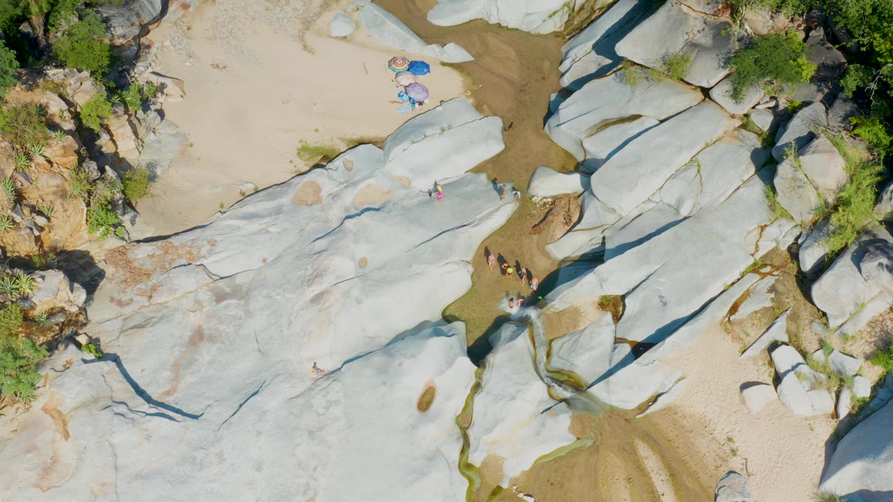 Aerial view of smooth white rock slabs, sand, shrubs, umbrellas, and people.