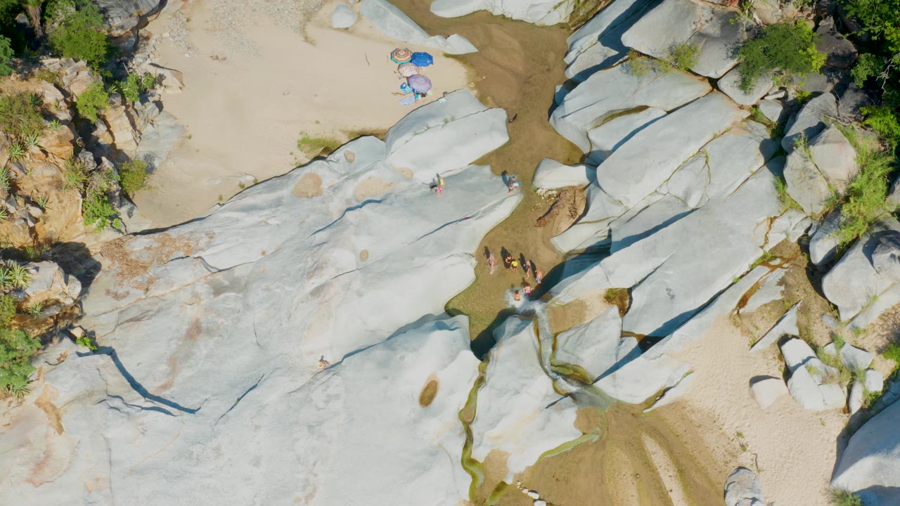 Top-down view in La Palma, Baja California Sur with pale boulders crossing the frame, a sandy wash at the top, shrubs to the side, and several small people on the rock.