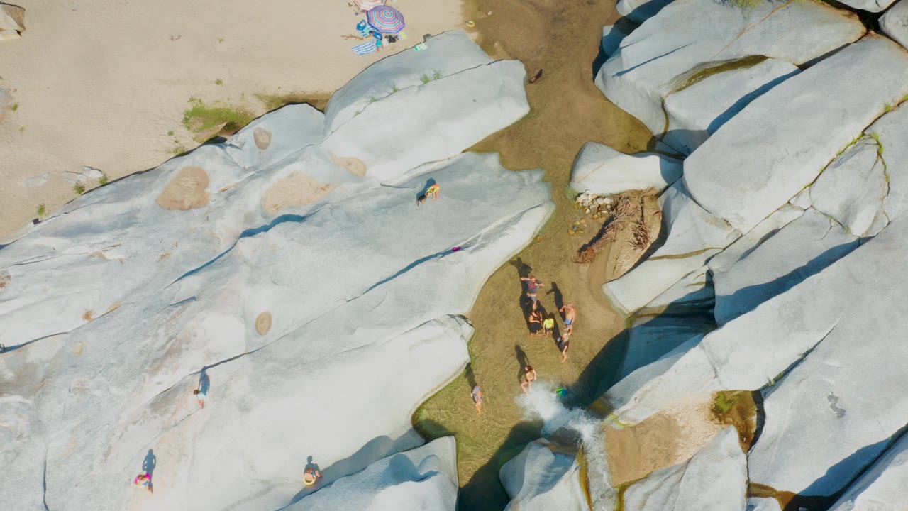 Aerial frame over La Palma's rocky beach edge showing pale stone plates, tan sand with colorful umbrellas, shallow water, and small visible people spread around the rocks.