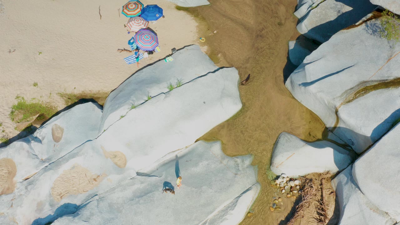Top-down aerial frame from La Palma, Baja California Sur showing four colorful umbrellas near pale sand, shallow water, white rock slabs, and a small person visible near the lower rocks.