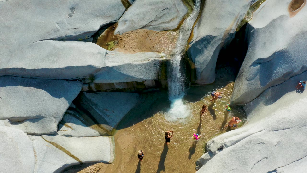 Drone view of friends standing and sitting near the waterfall pool.