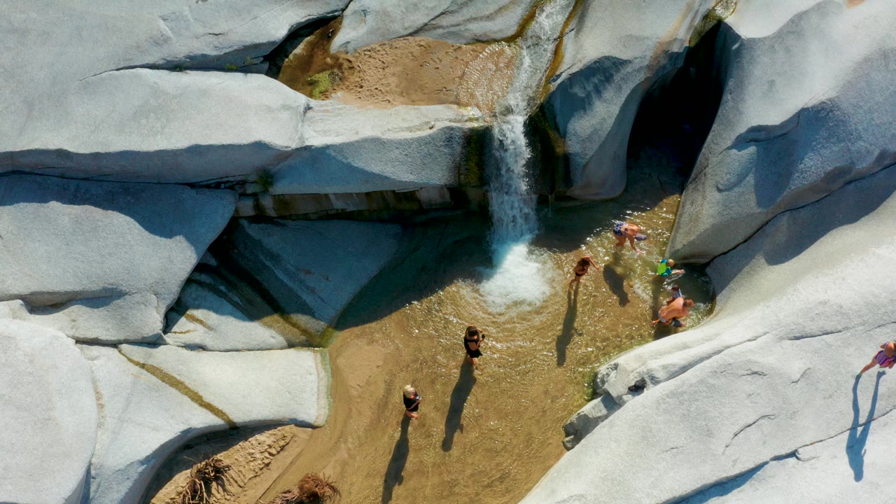 Overhead frame in La Palma, Baja California Sur showing children and adults beside pale rock slabs, shallow water, and a small waterfall entering a tan pool.