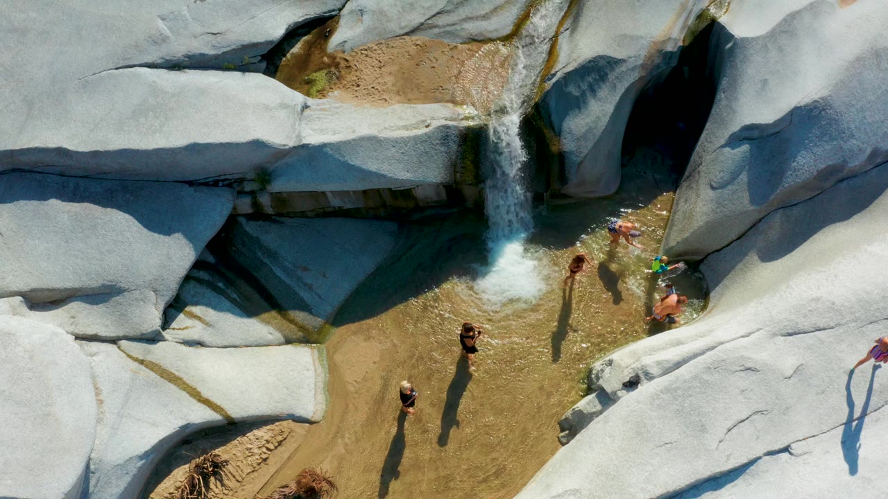 Children stand in shallow water below a small waterfall among smooth rocks.