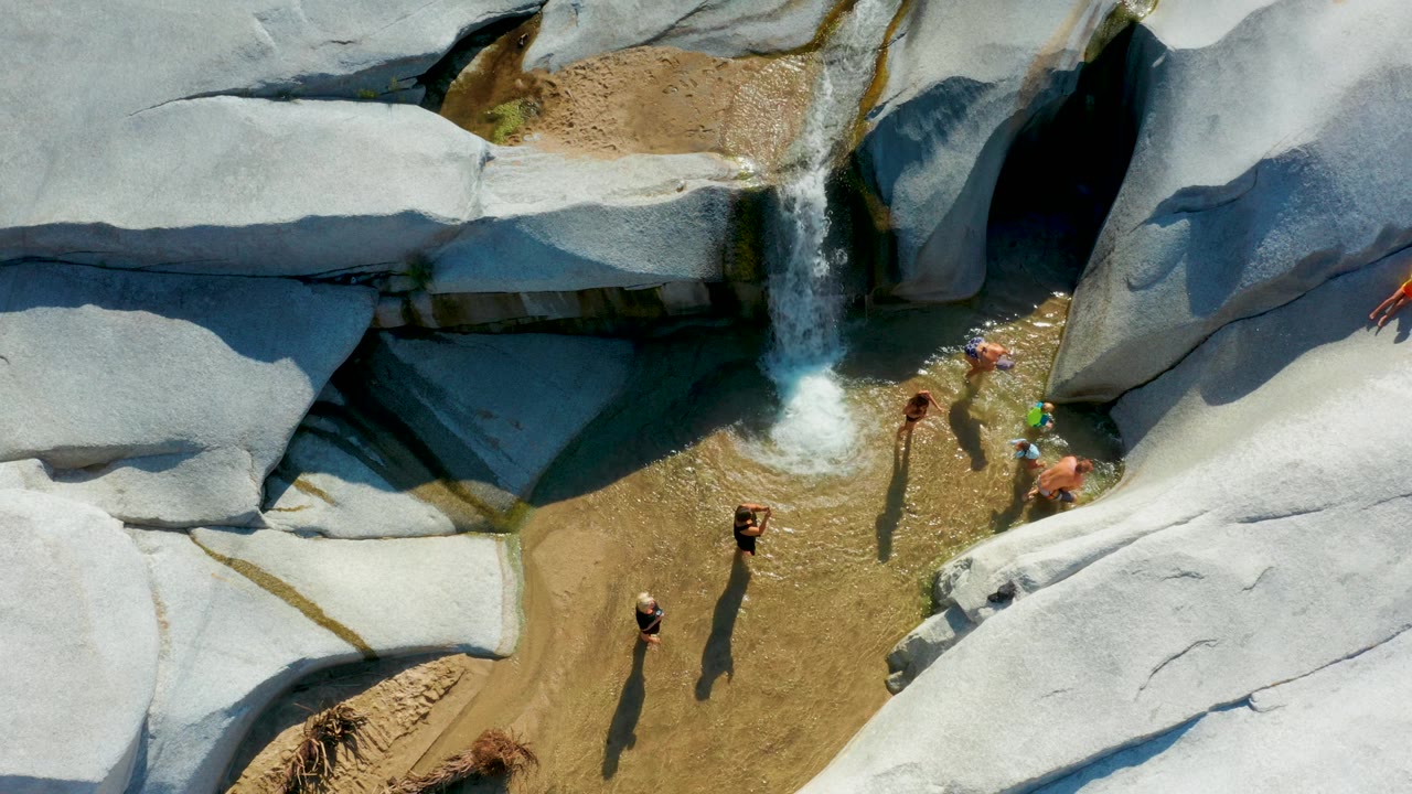 Overhead La Palma frame with a child or youth in bright clothing sliding across a smooth pale rock toward the shallow pool where other children stand near the falls.