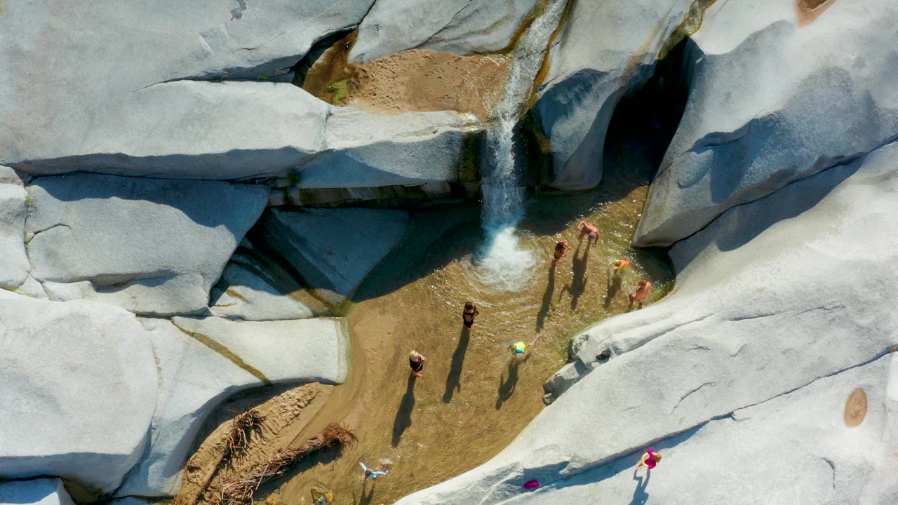 Overhead frame from La Palma, Baja California Sur showing several people in and beside shallow clear water below a small waterfall, with pale gray rocks around the pool.