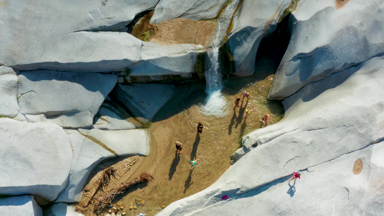 Top-down La Palma frame with people spaced across the shallow water and rock edge, a small waterfall feeding the pool, and pale stone surfaces dominating the frame.