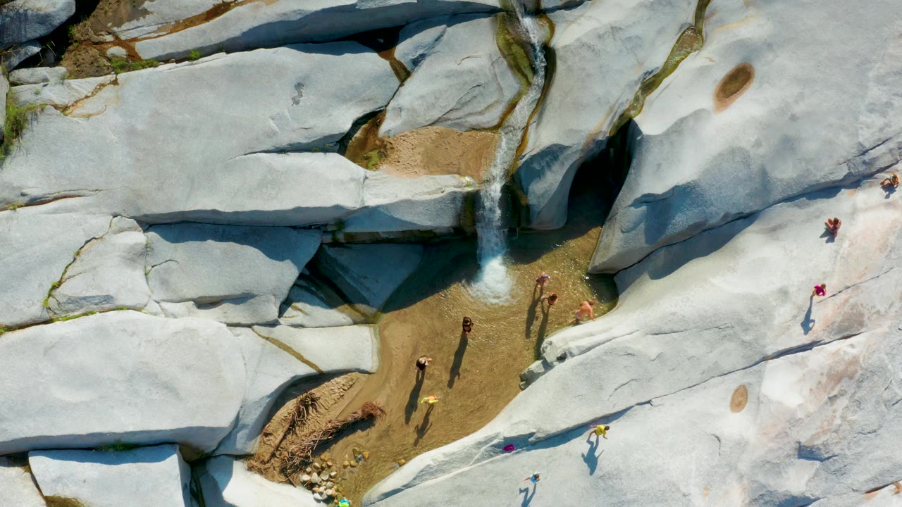 Drone view of a waterfall channel with hikers spread across large rock slabs.