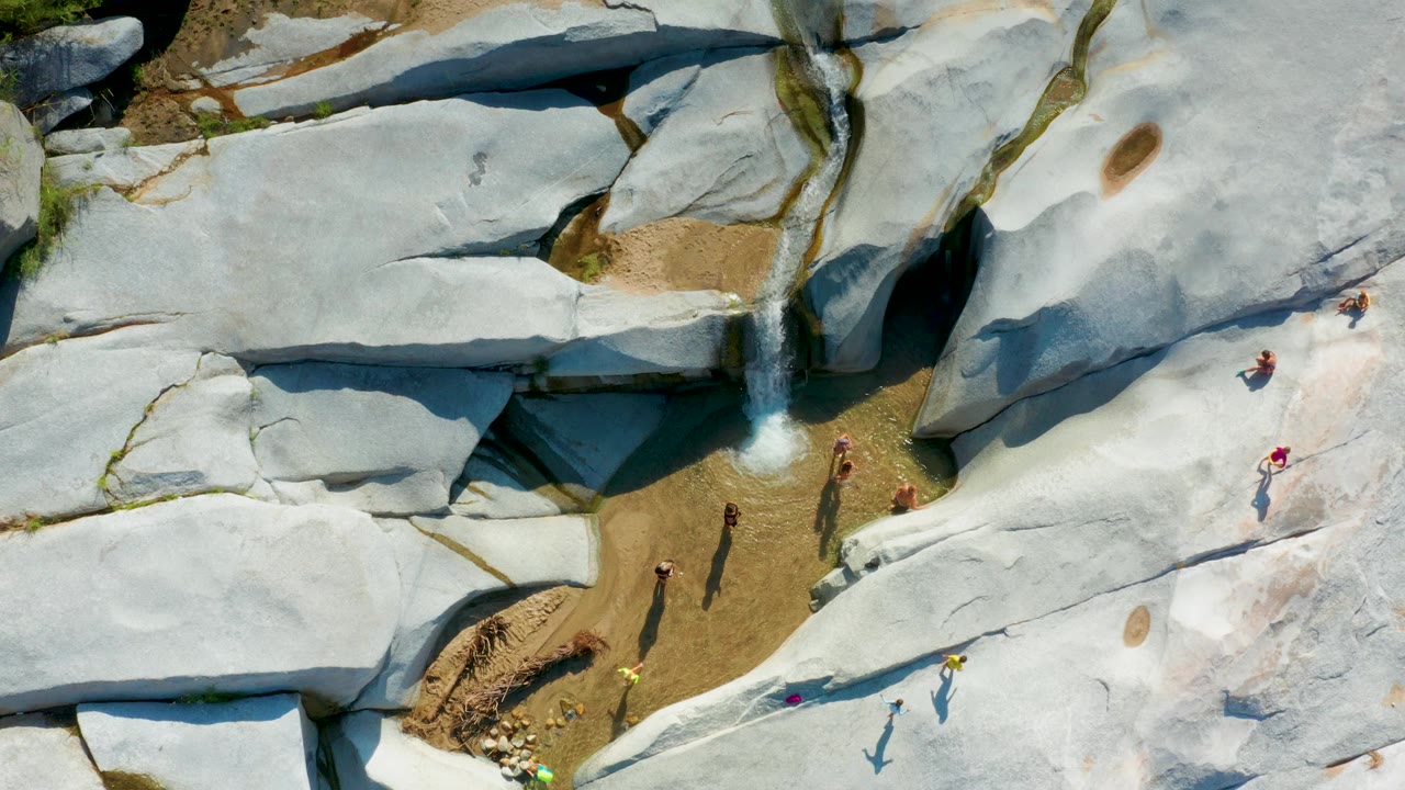 Top-down frame from La Palma, Baja California Sur showing a small waterfall at the left of the rock formation and people in bright clothing crossing the smooth pale stone.