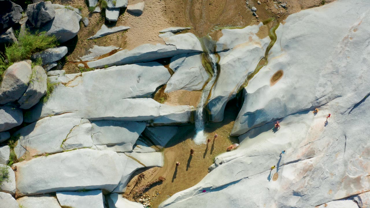Overhead drone-style frame in La Palma, Baja California Sur showing pale rock slabs, sandy patches, a narrow water channel, shrubs, and several small visible people near the pools.
