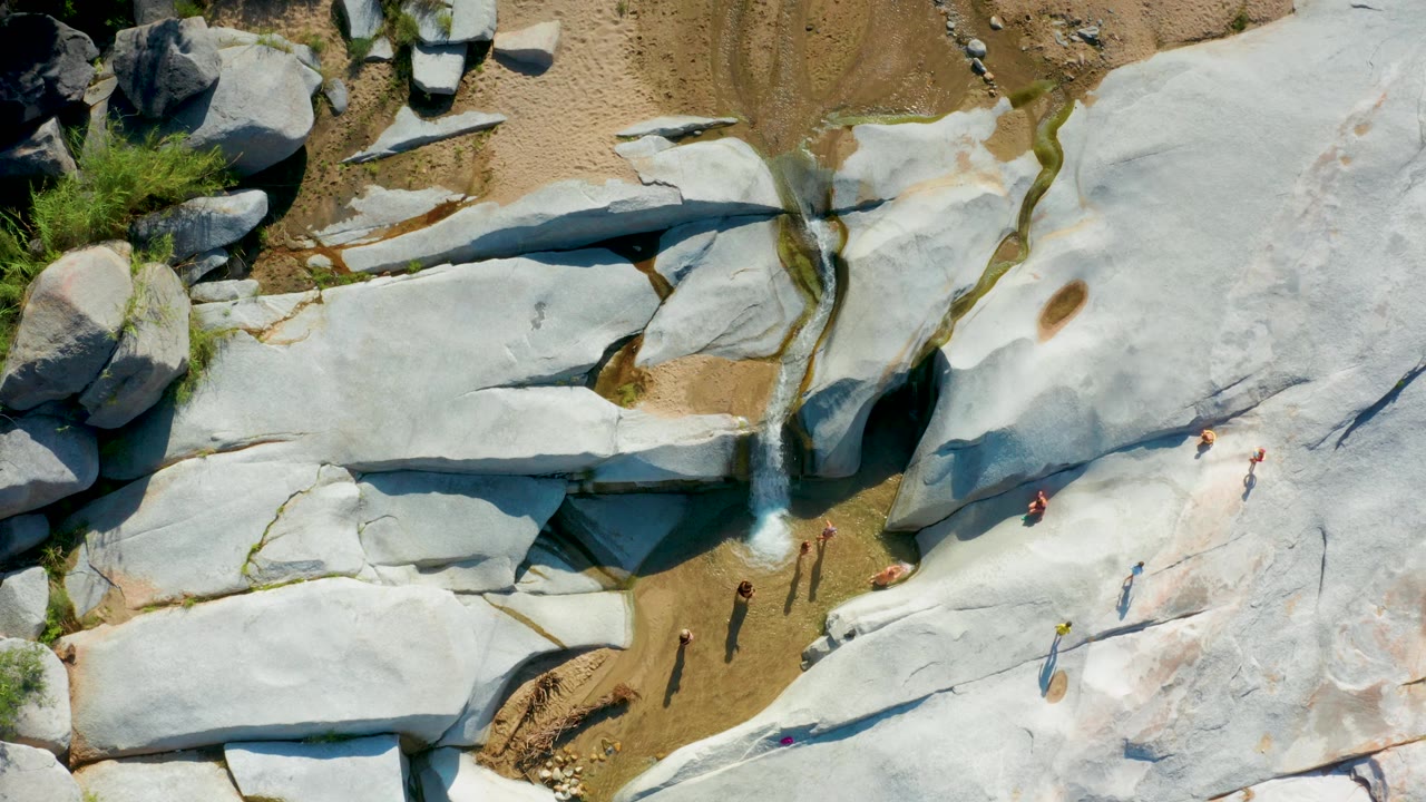 The frame shows pale rock shelves and a narrow stream in La Palma, Baja California Sur, with sandy ground above the rocks and small visible people spread across the sunlit surface.