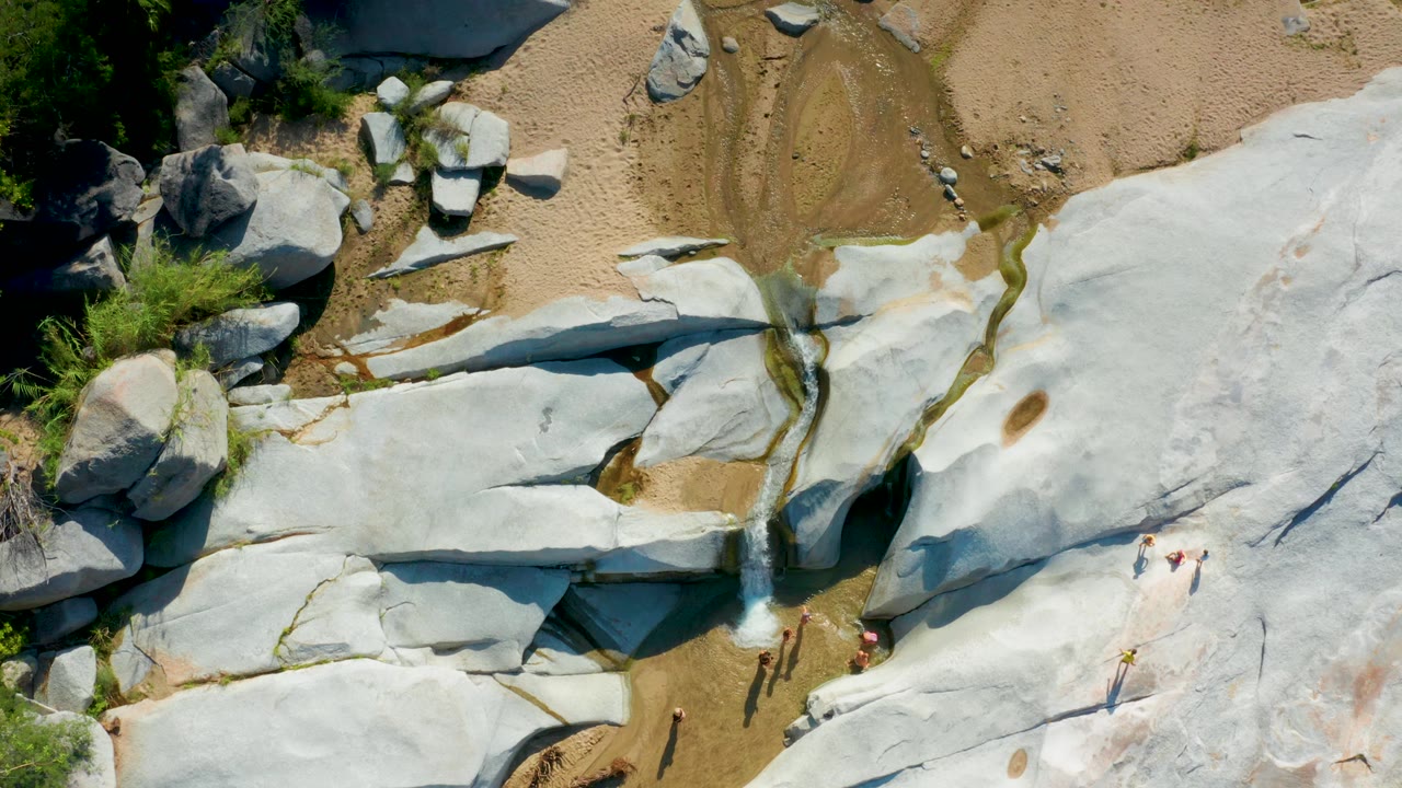 Aerial view of pale rock shelves, sandy water channels, greenery, boulders, and several small people on the rock.