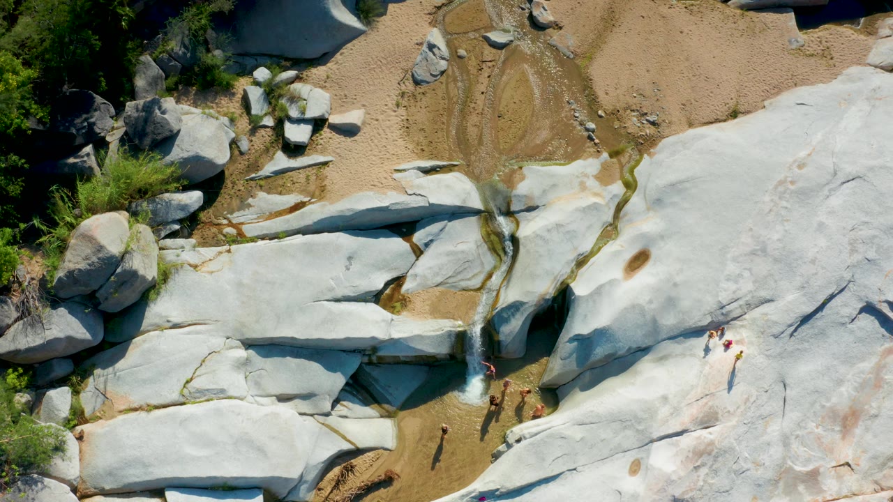 Top-down La Palma frame showing smooth white stone across the lower right, sandy channels at the top, green bushes on the left edge, and tiny people gathered beside the waterfall.