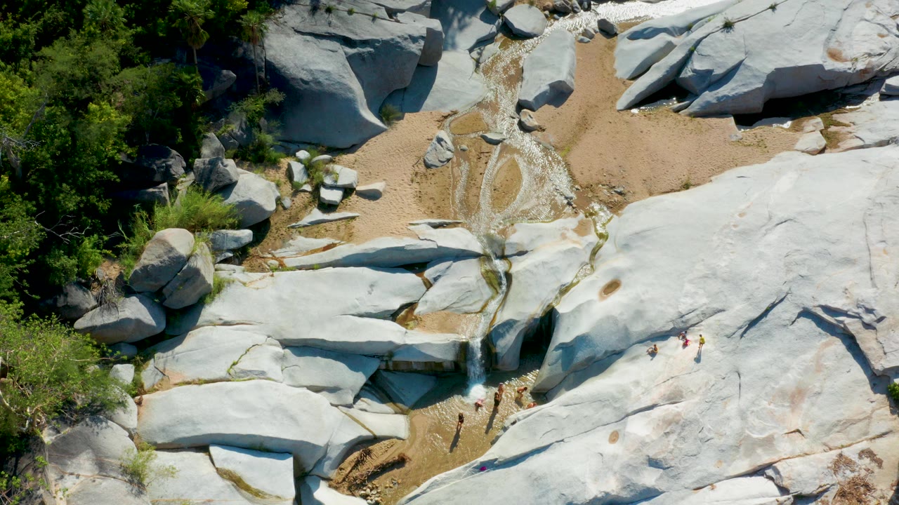Overhead view of pale rock slabs, sand, green vegetation, and a small water drop.