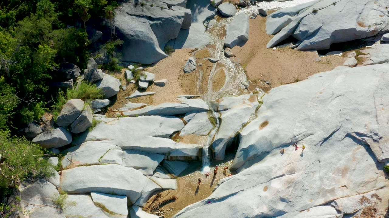 Aerial frame from La Palma, Baja California Sur with grey rock shelves, tan sand, scattered boulders, small water channels, surrounding green trees, and no visible people.