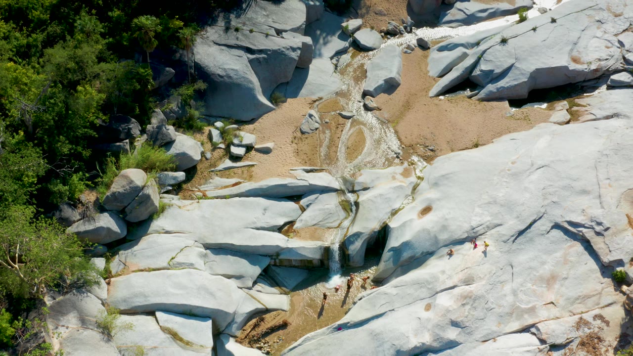Top-down La Palma frame showing pale rock sheets on the right, sandy wash through the middle, thin water streaks near the center, dense greenery on the left, and no visible people.
