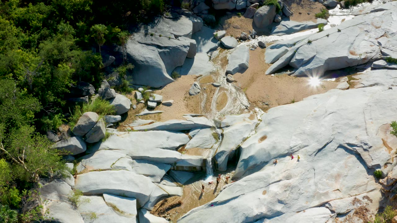 Overhead frame from La Palma, Baja California Sur where a bright sun glint flashes on the rock area beside sand, small water channels, surrounding trees, and no visible people.