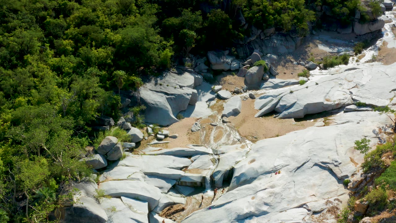 A slightly shifted aerial frame in La Palma, Baja California Sur, with a broad pale rock shelf, tan sediment, green canopy, and thin wet channels crossing the clearing, with no visible people.
