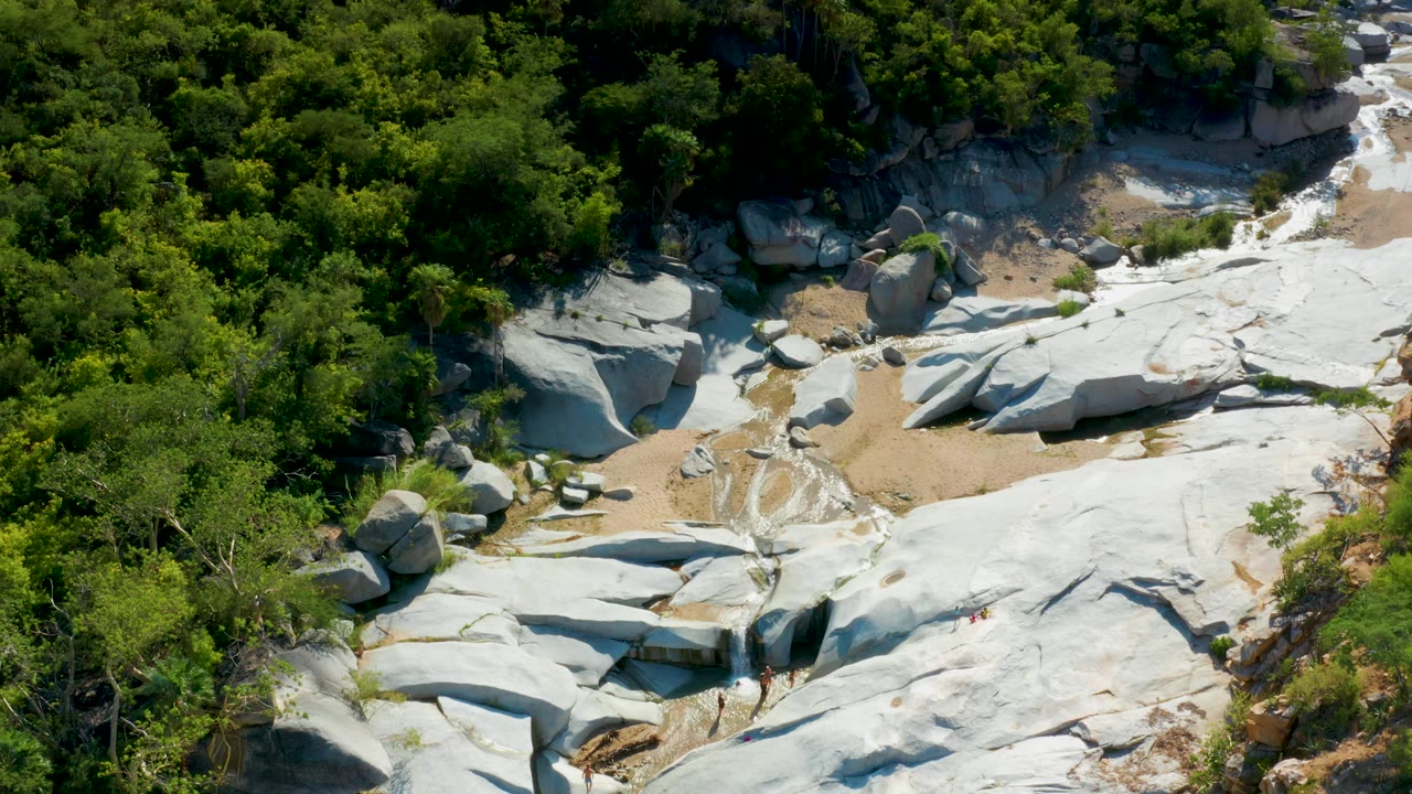 Aerial frame in La Palma, Baja California Sur, with large pale boulders near the forest edge, sand-colored ground between rocks, and thick green vegetation, with no visible people.