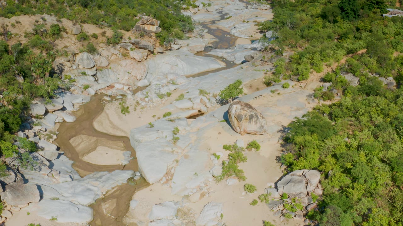 Aerial frame over La Palma, Baja California Sur, with broad light-colored rock sheets, a narrow damp channel, green vegetation along the edges, and no visible people.