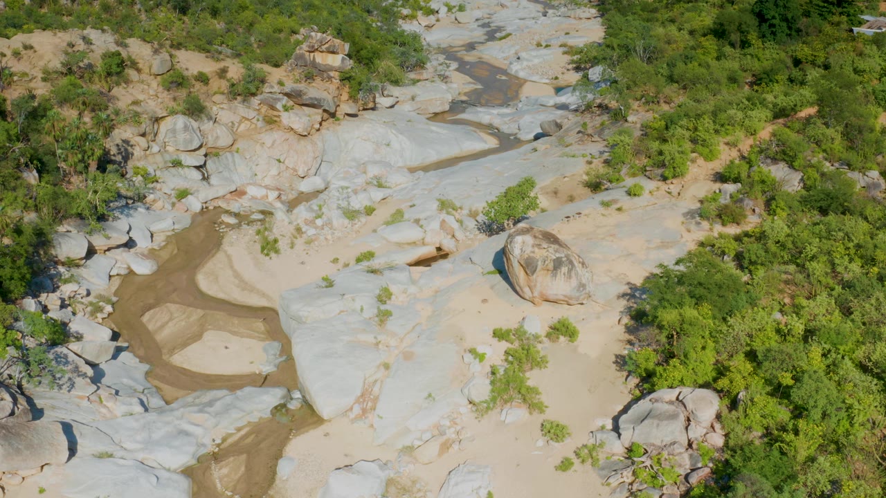 Mid-clip aerial frame from La Palma, Baja California Sur, showing smooth pale canyon rock, small sand pockets, rounded boulders, brush, and no visible people.