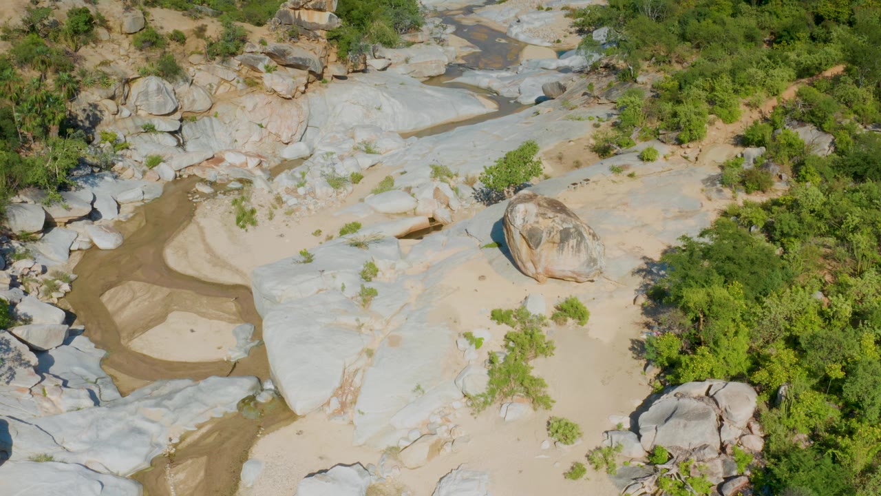 A high aerial frame over a rocky riverbed in La Palma, Baja California Sur, showing pale stone slabs, tan sand, a narrow muddy channel, scattered shrubs, and no visible people.